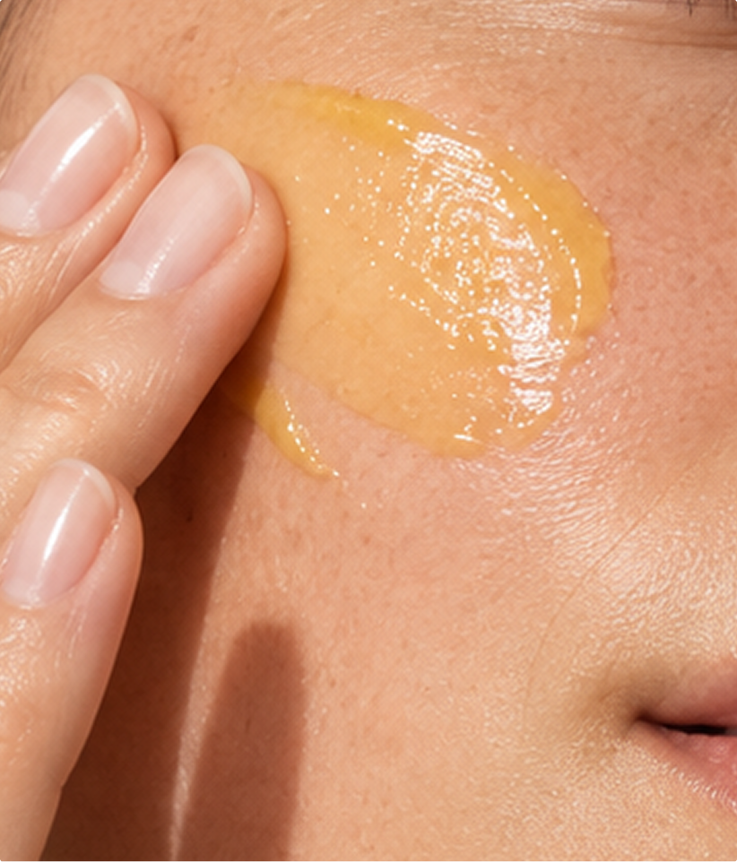 Close-up of a person's cheek with a glossy, translucent golden-yellow cosmetic cream or gel being spread across the skin by a hand with neatly manicured nails.