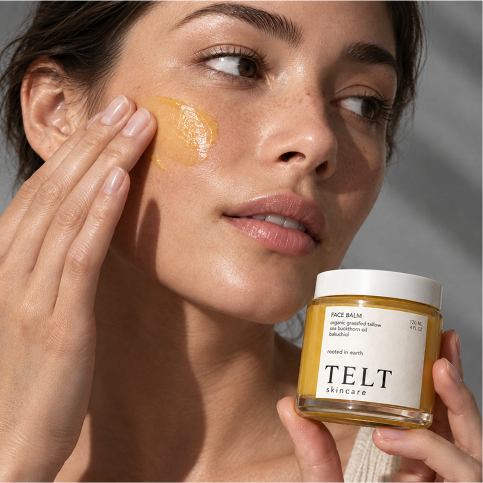 A close-up lifestyle shot of a woman applying a golden-yellow face balm to her cheek while holding a jar of TELT Skincare Face Balm in her other hand.